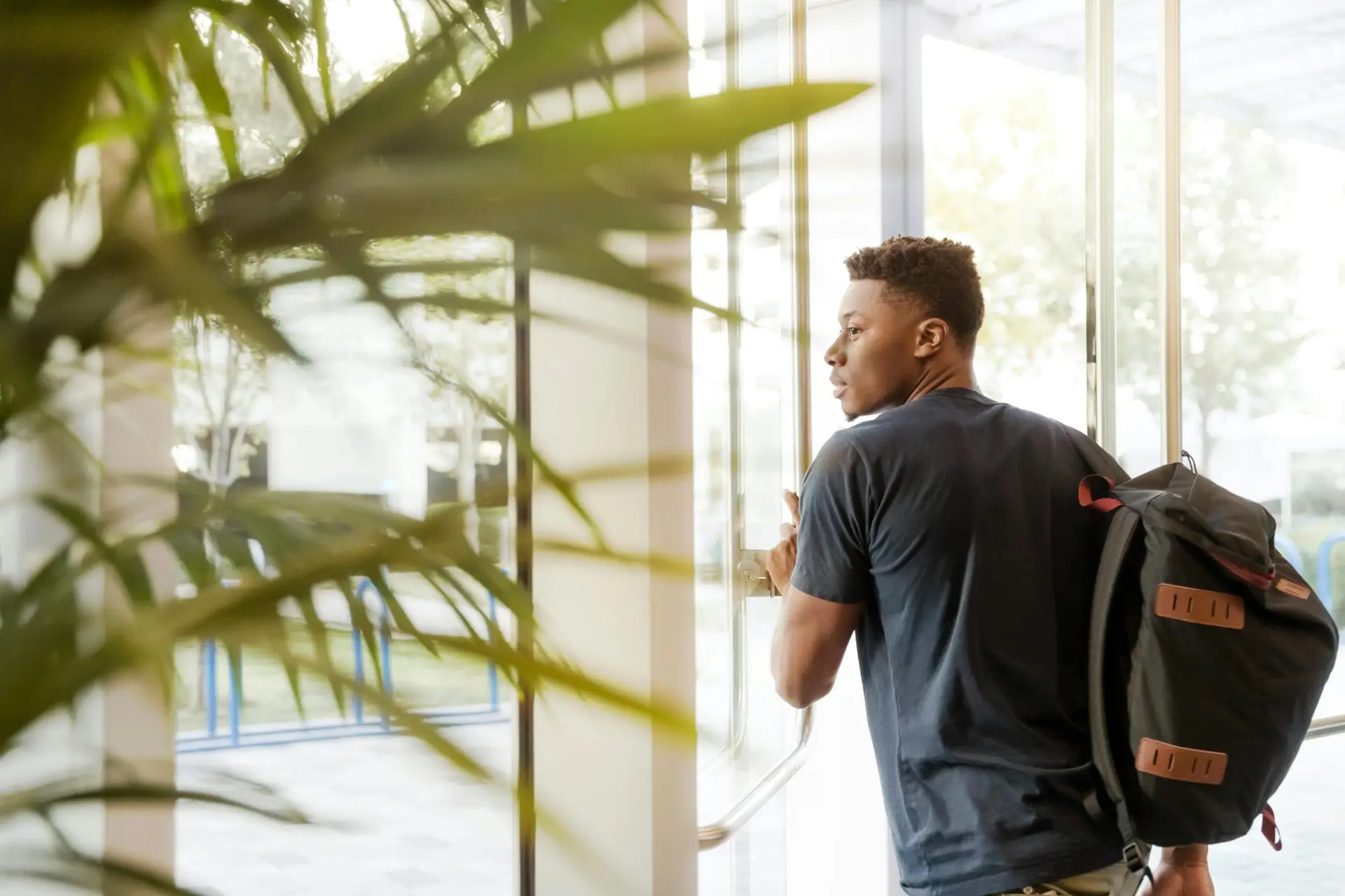 A college student with a backpack exits a campus building through glass doors.